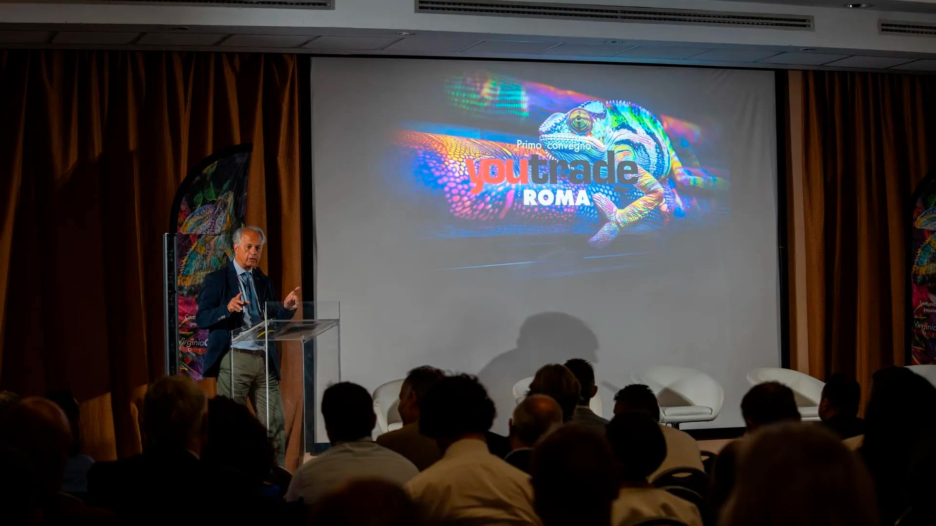 Man in a suit delivering a presentation at a conference, standing behind a clear lectern while a colorful lizard graphic and the text 'youtrade ROMA' appear on a large projection screen behind him.