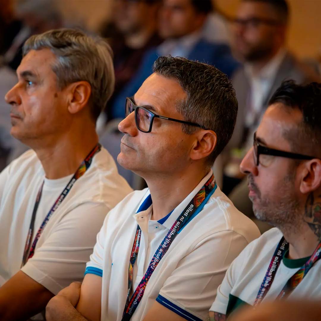 Three men in the audience at a conference, wearing white polo shirts and colorful lanyards, listening intently.